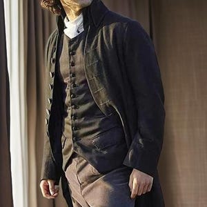 May include: A dark, long coat with a high collar, worn over a buttoned vest and trousers. The coat has multiple buttons down the front and long sleeves. The vest is also buttoned and has pockets. The trousers are a muted color.