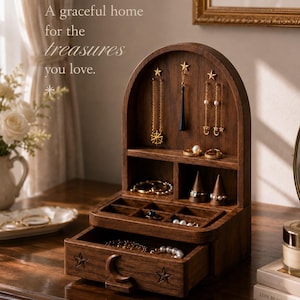 May include: A dark wood jewelry organizer with an arched top, featuring hooks for necklaces, ring cones, small compartments, and a pull-out drawer with carved star and moon motifs. The text "A graceful home for the treasures you love." is visible.