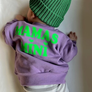Op de afbeelding: Een kind draagt een paarse trui met de woorden "MAMAS MINI" in felgroen. Het kind draagt ook een groene gebreide muts. De onderkant van het kind draagt een grijs-witte gestreepte broek.