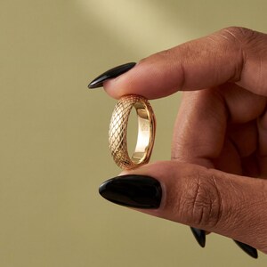 May include: A gold ring featuring a textured, scale-like pattern on the exterior surface. The band has a smooth, polished interior finish with a small, clear hallmark stamp visible on the inside of the metal band, showcasing a detailed design.