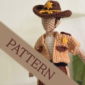 Puede incluir: Muñeca de ganchillo con sombrero de vaquero marrón con detalles amarillos, chaqueta beige y pantalones marrones. La muñeca tiene un tono de piel neutro y una camisa blanca. La palabra "PATTERN" está impresa en diagonal en la imagen.