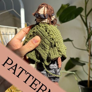 May include: A handmade crochet doll with brown hair, a green cape, and gray pants. The doll has a beige face and is wearing a hat. The word "PATTERN" is printed on a diagonal banner across the image. The doll is held in a hand.