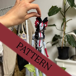 Puede incluir: Muñeca de ganchillo hecha a mano con un diseño tribal. La muñeca presenta un cuerpo azul claro, detalles en rojo y negro y pelo largo negro. La palabra "PATTERN" está impresa en la imagen en una pancarta roja.