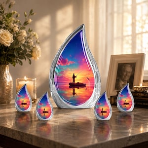 May include: A silver-colored teardrop-shaped memorial urn with a sunset scene of a person fishing on a boat. Four smaller teardrop-shaped urns are also present. The urns are on a marble-like surface with a vase of flowers and a framed photo in the background.