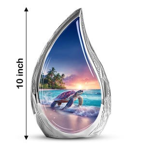 May include: A decorative silver-coloured teardrop-shaped sculpture. The interior features a vibrant beach scene with a sea turtle, ocean waves, and a sunset. The sculpture is 25.4 cm tall.