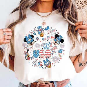 May include: A cream-colored t-shirt featuring a circular graphic print with the text USA in red, white, and blue. The design includes cartoon characters wearing patriotic hats and glasses, fireworks, stars, and various American-themed icons.