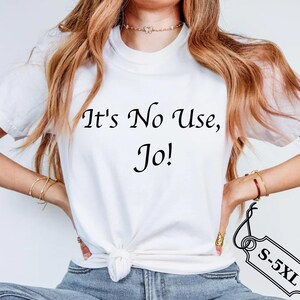 May include: White t-shirt featuring the text "It's No Use, Jo!" in black script. The shirt is knotted at the waist. A size tag, S-5XL, is attached. The model is wearing blue jeans.
