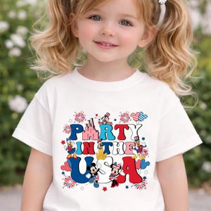 May include: A white short-sleeved t-shirt featuring a colorful graphic print with the text PARTY IN THE USA. The design includes illustrations of Mickey Mouse, Minnie Mouse, Donald Duck, Daisy Duck, Pluto, and Goofy, accented with fireworks and stars.