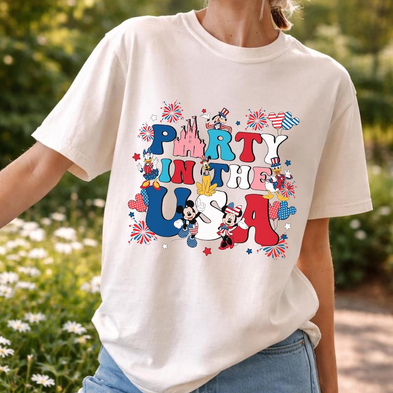 May include: A cream-colored t-shirt featuring a graphic print with the text PARTY IN THE USA. The design includes illustrations of Mickey Mouse, Minnie Mouse, Donald Duck, Daisy Duck, and Pluto wearing patriotic attire, alongside a pink castle.