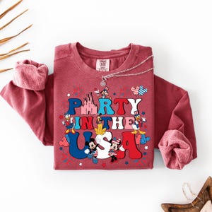 May include: A maroon long-sleeve crewneck sweatshirt featuring a graphic print with the text PARTY IN THE USA. The design includes cartoon characters, a castle, fireworks, and star shapes in red, white, and blue colors. A silver chain necklace rests on the collar.