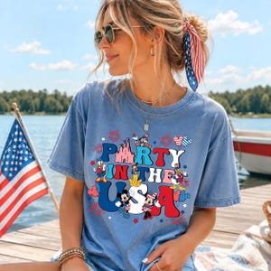 May include: A light blue short-sleeve t-shirt featuring a graphic print with the text PARTY IN THE USA, decorated with red, white, and blue fireworks, balloons, and cartoon characters wearing patriotic hats and outfits in a festive style.