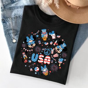 May include: A black t-shirt featuring a circular graphic print with the text USA in red, white, and blue. The design includes cartoon characters wearing patriotic accessories, fireworks, stars, and American flag patterns in red, white, and blue.