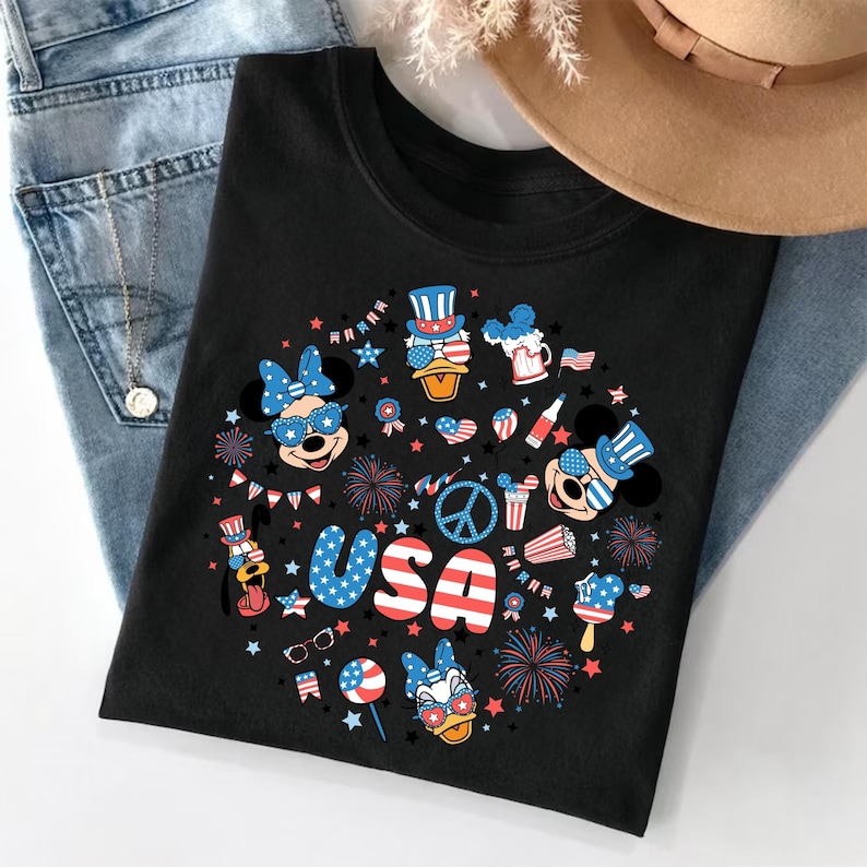 May include: A black t-shirt featuring a circular graphic print with the text USA in red, white, and blue. The design includes cartoon characters wearing patriotic accessories, fireworks, stars, and American flag patterns in red, white, and blue.