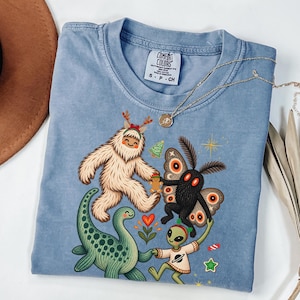 May include: A blue crew neck t-shirt featuring a graphic print of a white yeti, a black moth, a green dinosaur, and a green alien. The yeti wears a red bow, the alien wears a shirt with a planet icon, and small holiday icons surround them.