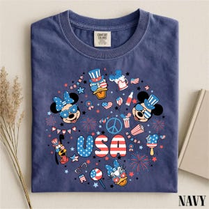 May include: A navy blue t-shirt featuring a circular graphic print with the text USA in red, white, and blue. The design includes cartoon characters wearing patriotic hats and sunglasses, fireworks, stars, and American flag-themed icons.