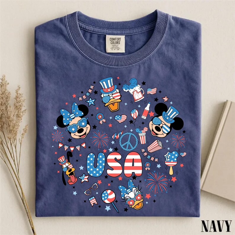 May include: A navy blue t-shirt featuring a circular graphic print with the text USA in red, white, and blue. The design includes cartoon characters wearing patriotic hats and sunglasses, fireworks, stars, and American flag-themed icons.