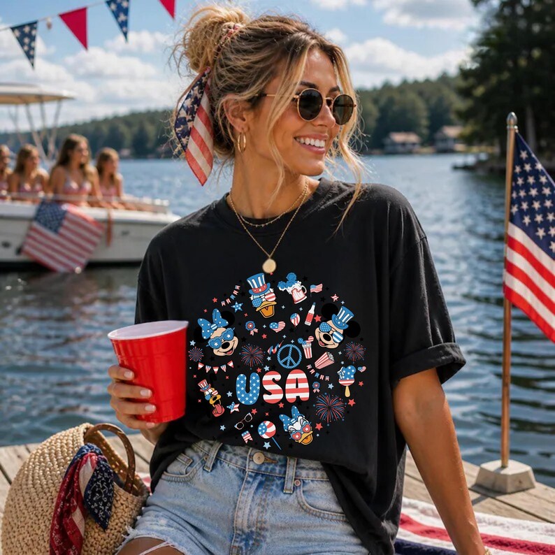 May include: A black short-sleeved t-shirt featuring a circular graphic print with the text USA in red, white, and blue. The design includes cartoon character heads wearing patriotic hats, fireworks, a peace sign, and various red, white, and blue icons.