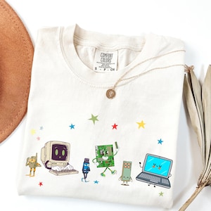 May include: A cream-colored t-shirt featuring a graphic print of anthropomorphic computer components, including a vintage monitor, a USB drive, a circuit board, and a laptop, surrounded by small, colorful stars. The shirt has a Comfort Colors label.