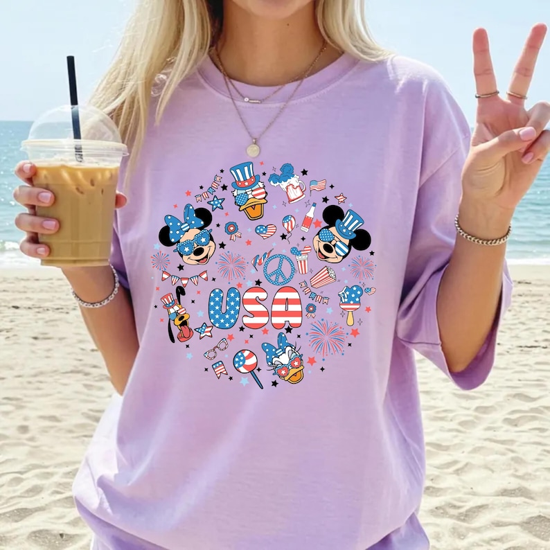 May include: A light purple t-shirt featuring a circular graphic print with the text USA in red, white, and blue. The design includes cartoon characters wearing patriotic hats and sunglasses, fireworks, stars, and various American-themed icons.