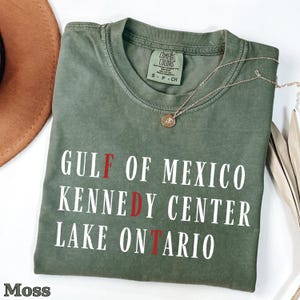 May include: A moss green short-sleeve t-shirt with a crew neckline. The shirt features white text that reads GULF OF MEXICO, KENNEDY CENTER, and LAKE ONTARIO, with the letters F, D, and T highlighted in red. A small brand label is at the neck.