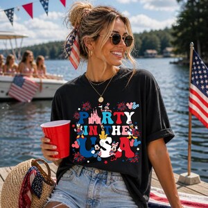 May include: A black short-sleeved t-shirt featuring a graphic print with the text PARTY IN THE USA, a pink castle silhouette, and cartoon characters dressed in red, white, and blue patriotic outfits, surrounded by firework and star motifs.