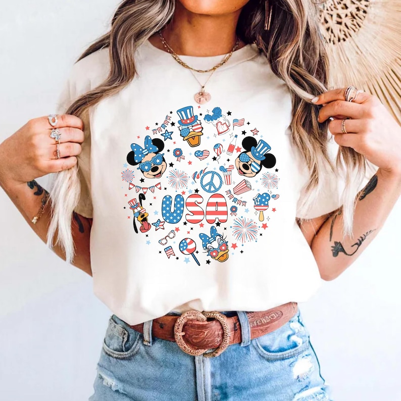 May include: A cream-colored t-shirt featuring a circular graphic print with the text USA in red, white, and blue. The design includes cartoon characters wearing patriotic hats and glasses, fireworks, stars, and various American-themed icons.