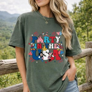May include: A dark olive green t-shirt featuring a graphic print with the text PARTY IN THE USA. The design includes illustrations of various cartoon characters, a castle, and patriotic red, white, and blue star and firework accents.