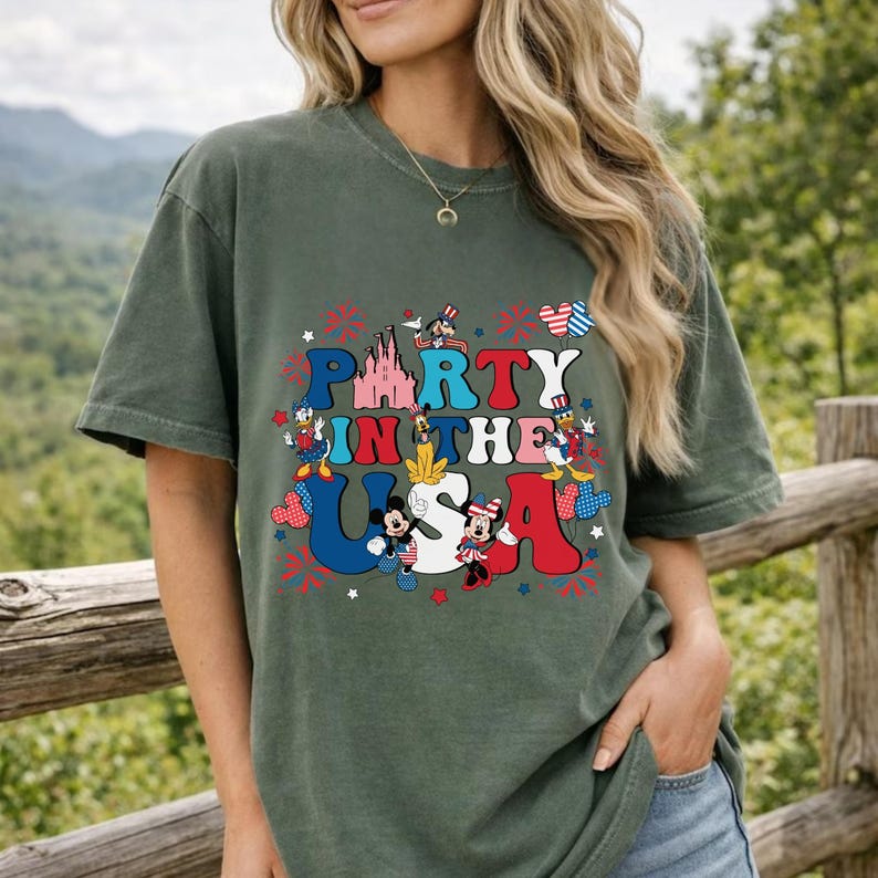 May include: A dark olive green t-shirt featuring a graphic print with the text PARTY IN THE USA. The design includes illustrations of various cartoon characters, a castle, and patriotic red, white, and blue star and firework accents.