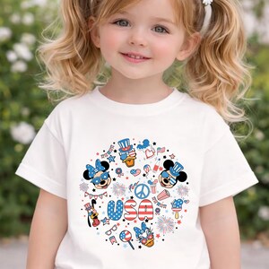 May include: A white short-sleeved t-shirt featuring a circular graphic print with red, white, and blue patriotic motifs. The design includes cartoon characters wearing star-spangled accessories, fireworks, a peace sign, and the text USA in bold letters.