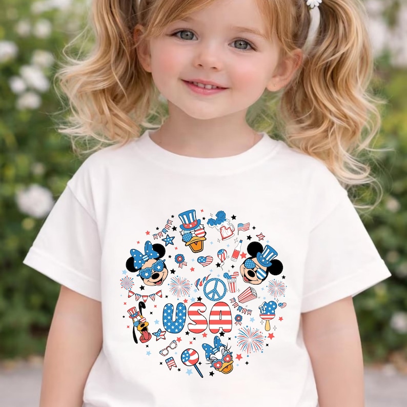 May include: A white short-sleeved t-shirt featuring a circular graphic print with red, white, and blue patriotic motifs. The design includes cartoon characters wearing star-spangled accessories, fireworks, a peace sign, and the text USA in bold letters.