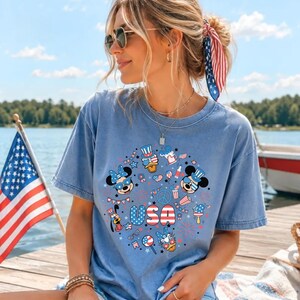 May include: A light blue short-sleeve t-shirt featuring a circular graphic print with cartoon characters wearing patriotic hats and sunglasses, surrounded by stars, fireworks, and the text USA in red, white, and blue stripes.