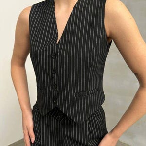 May include: Black and white pinstripe vest with a V-neck and button closure. The vest has a tailored fit and a small pocket on the front. The matching pants are also visible. This is a classic, professional outfit.