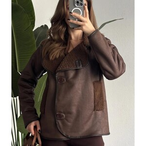 Puede incluir: Chaqueta marrón de piel sintética con cuello y ribete de borreguillo. La chaqueta tiene dos botones y un bolsillo en la parte delantera. La modelo lleva un bolso marrón. Una elegante opción de abrigo.
