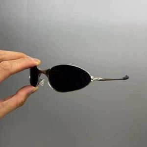 Może przedstawiać: Bezramkowe okulary przeciwsłoneczne z ciemnymi soczewkami. Oprawki są wykonane ze srebrnego metalu z brązowymi detalami na zausznikach. Okulary przeciwsłoneczne są trzymane na szarym tle.