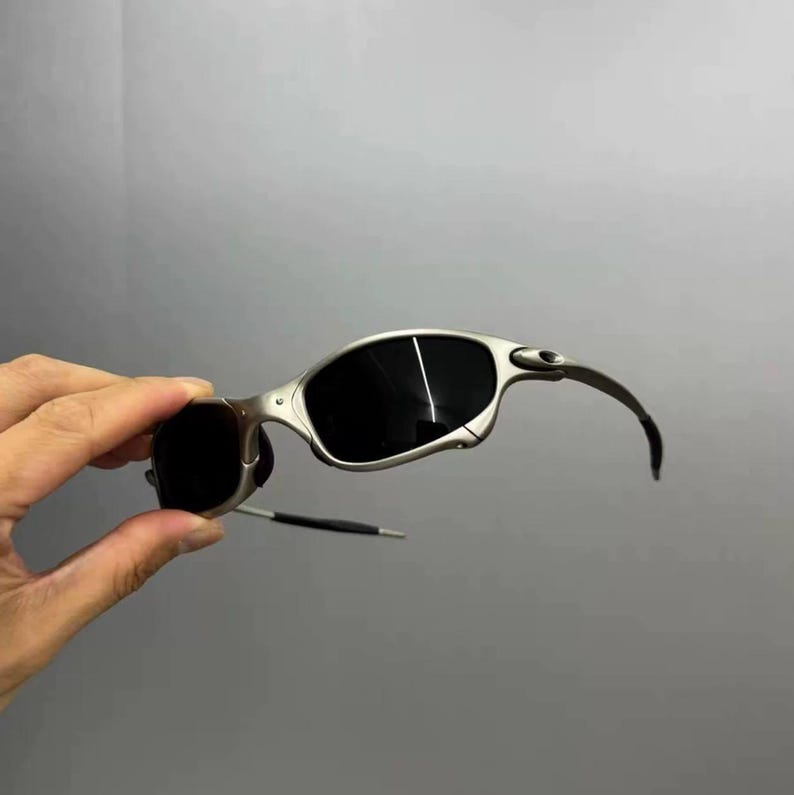 May include: Silver-framed sunglasses with dark lenses. The sunglasses have a curved design and a sleek, modern aesthetic. The lenses are a deep black, and the frame is a metallic silver color.