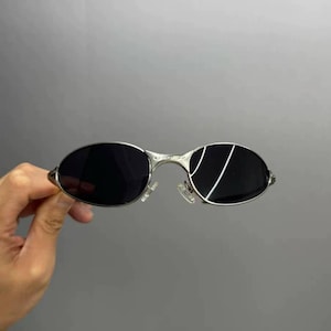 Może przedstawiać: Okulary przeciwsłoneczne w srebrnej oprawie z owalnymi czarnymi soczewkami. Okulary przeciwsłoneczne mają elegancki, nowoczesny design z cienką metalową oprawą i przezroczystymi noskami. Soczewki odbijają białą linię. Okulary są trzymane przez dłoń.