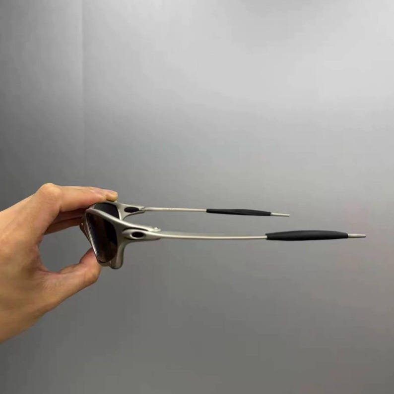 May include: Silver-framed sunglasses with dark lenses. The sunglasses have a sleek, modern design with thin temples and black earpieces. The frame is held by a hand against a gray background.
