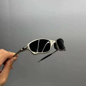 May include: Silver-framed sunglasses with dark lenses. The sunglasses have a sleek, modern design with a curved frame and a thin, straight arm. The lenses are black and reflect light. The sunglasses are held by a hand.