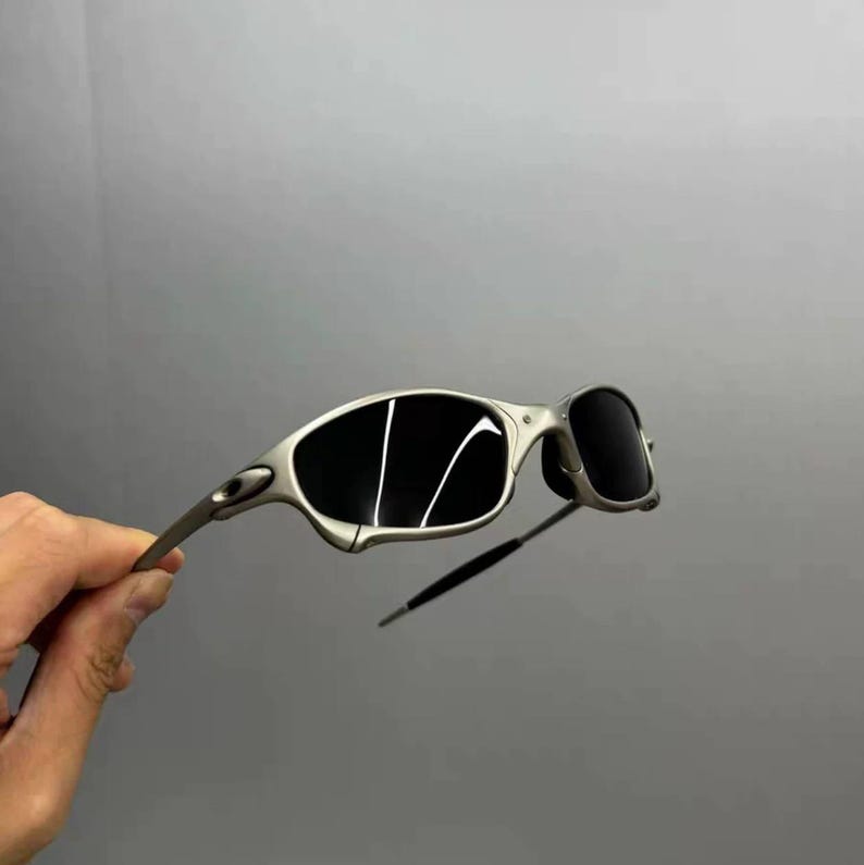 May include: Silver-framed sunglasses with dark lenses. The sunglasses have a sleek, modern design with a curved frame and a thin, straight arm. The lenses are black and reflect light. The sunglasses are held by a hand.