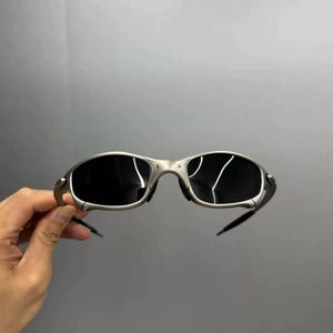 May include: Silver-framed sunglasses with dark lenses. The sunglasses have a sleek, modern design with a curved frame and a minimalist aesthetic. The lenses are dark, providing shade and protection from the sun.