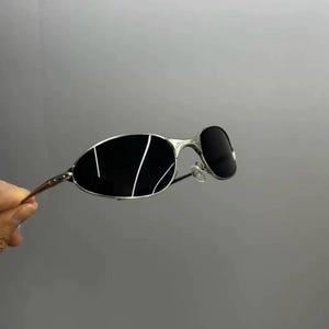 Może przedstawiać: Srebrne okulary przeciwsłoneczne z czarnymi soczewkami i białymi liniami akcentującymi. Okulary mają zakrzywioną konstrukcję. Obraz przedstawia okulary na szarym tle.
