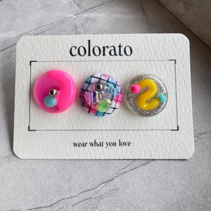 以下が含まれることがあります： coloratoおよびwear what you loveと書かれた白い台紙に固定された3つの円形スタッドピアス。左は銀色のビーズが付いた鮮やかなピンク色。中央は黒い格子模様に虹色のアクセント。右は銀色のラメ地に黄色い波線と2つの小さなビーズがあしらわれています。