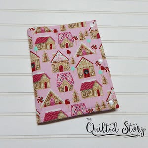 May include: A pink fabric cover with a repeating pattern of gingerbread houses, trees, and candy canes. The cover has white snaps and the text "The Quilted Story" in black script.