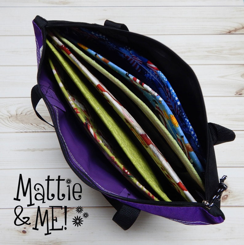 May include: A black tote bag with a purple interior, partially unzipped to reveal several colorful fabric panels. The panels feature various patterns, including floral and patriotic designs. The text "Mattie & ME!" is visible.