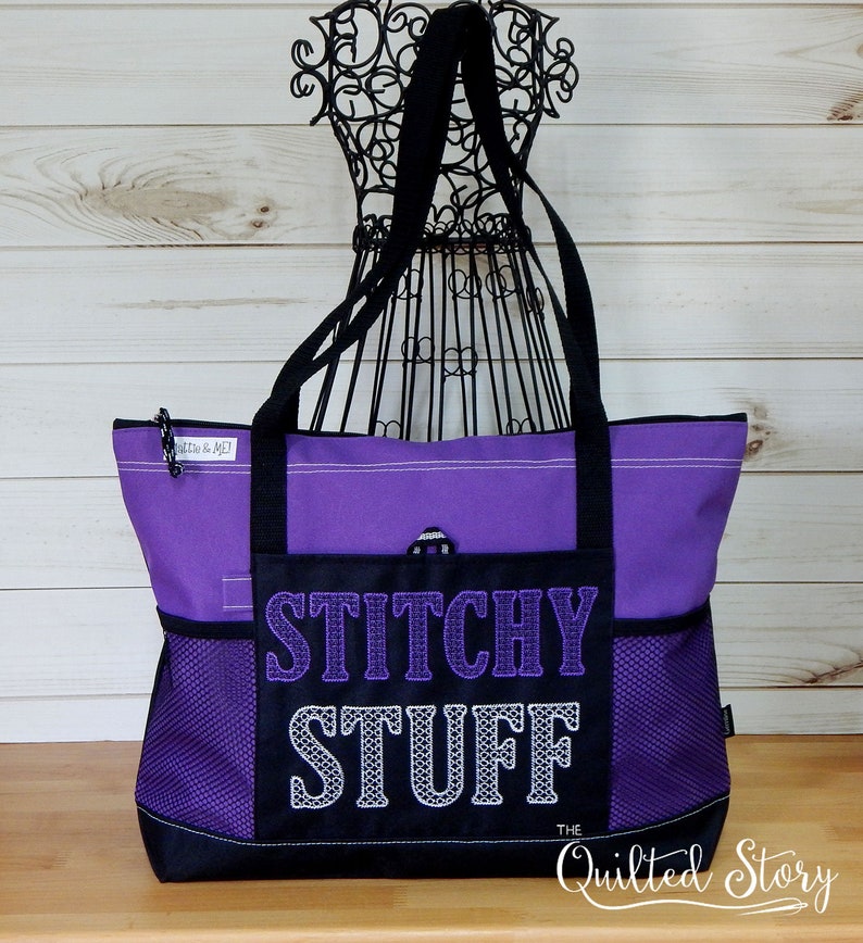 May include: A purple tote bag with black accents and handles. The front pocket features the embroidered text "STITCHY STUFF" in purple and white. The bag has mesh side pockets and a zipper closure. The words "The Quilted Story" are at the bottom.