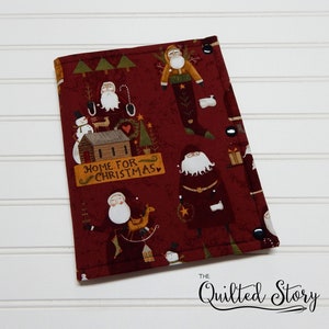May include: A burgundy fabric item with a Christmas-themed pattern featuring Santa Claus figures, snowmen, and the text "HOME FOR CHRISTMAS." The item is secured with black snaps and has a white background.