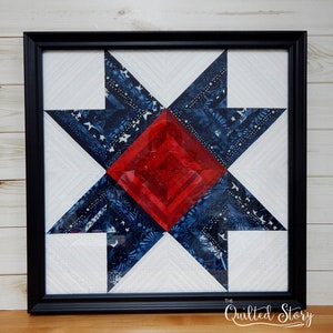 May include: A framed quilt block with a red, white, and blue star design. The star is made of fabric with a blue and white pattern. The quilt block is framed in a black frame. The text "The Quilted Story" is in the bottom right corner of the image.