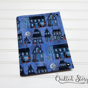 May include: A rectangular fabric cover featuring a blue background with a repeating pattern of dark houses, bare trees, full moons, and small orange pumpkins. The cover is secured with three black snaps along the right edge and includes the text The Quilted Story.