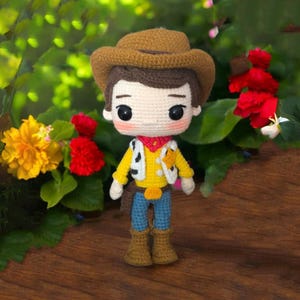 Może przedstawiać: Ręcznie robiona szydełkowa lalka przedstawiająca postać kowboja, z brązowym kapeluszem, żółtą koszulą, niebieskimi spodniami i brązowymi butami. Lalka ma białą kamizelkę z czarnymi kropkami i czerwoną chustą. Lalka jest ustawiona na tle zielonych liści i kolorowych kwiatów.