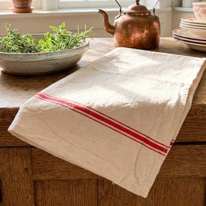 May include: A rectangular off-white kitchen towel featuring two parallel red stripes running horizontally across the fabric. The towel is folded and placed on a wooden surface, showcasing a simple woven texture and clean, straight edges.